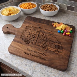 May include: A walnut cutting board with a handle, engraved with a detailed outline of a recreational vehicle. The text "The Jensen Family's Road Trip Fuel Station" is below the RV. The board measures 26.67 x 40.64 cm.