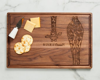 May include: A wooden cutting board with a giraffe design engraved on it. The board has the text "Did someone say Cheese?!" engraved on it. There are crackers and cheese on the board.