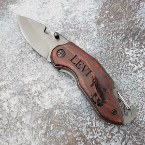 May include: A personalised pocket knife with a wooden handle. The handle is engraved with the name "Levi" and a fish design. The knife has a silver blade and a bottle opener.