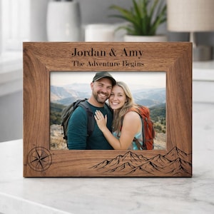 Peut inclure: Un cadre photo en bois avec les noms "Jordan & Amy" et la phrase "The Adventure Begins" gravés. Le cadre présente une boussole et un motif de montagnes. Il contient une photo d'un couple souriant.