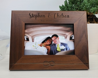 May include: A personalized wooden picture frame with a dark brown finish. The frame has an engraved inscription that reads "Stephen & Chelsea Est. 07.07.2022" and a decorative knot design. The frame holds a photo of a couple in a car.