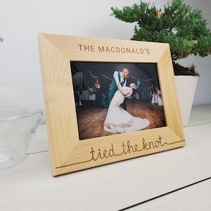 May include: A light-coloured wooden picture frame with the words "THE MACDONALD'S" and "tied the knot" engraved on it. The frame holds a photo of a couple dancing at a wedding. A small green plant is in the background.