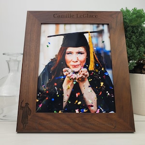 Peut inclure: Un cadre photo en bois brun foncé avec une photo de diplômé d'une personne portant une toque et une robe noires. Le cadre est gravé avec le texte "Camille LeGlace", "Masters of Education" et "Waterloo University".