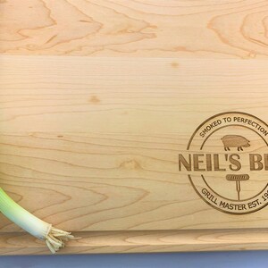 May include: A light wood cutting board with a round, engraved logo that says "Smoked to Perfection Neil's BBQ Grill Master Est. 1962" and a small pig illustration.