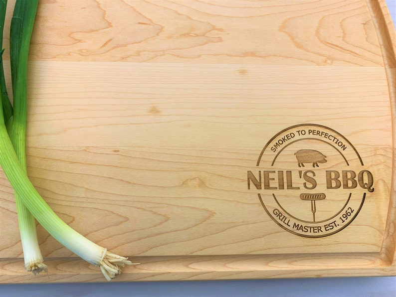 May include: A light wood cutting board with a round, engraved logo that says "Smoked to Perfection Neil's BBQ Grill Master Est. 1962" and a small pig illustration.