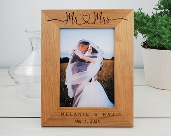 May include: A wooden picture frame with a photo of a couple in a field. The frame is engraved with "Mr & Mrs" and the couple's names and wedding date: "Melanie & Raj, May 1, 2024".