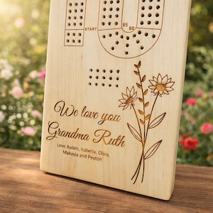 May include: Wooden cribbage board with engraved game layout, holes, and floral design. The board includes the text "We love you Grandma Ruth" and names. A thoughtful gift for family.