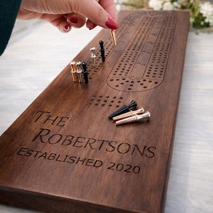 May include: A wooden cribbage board with engraved text reading "THE ROBERTSONS ESTABLISHED 2020." The board has a series of holes for pegs, with a hand placing a gold peg. Additional pegs in gold, silver, and black are also visible.