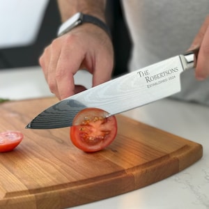 May include: A person is using a silver kitchen knife with a wooden handle to cut a red tomato on a wooden chopping board. The knife has the text "THE ROBERTSONS ESTABLISHED 2024" engraved on the blade.