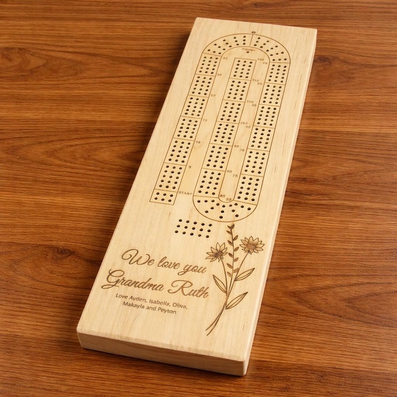 May include: A wooden cribbage board with a classic track design. The board is engraved with the text "We love you Grandma Ruth" and the names of the people who love her. A floral design is also engraved on the board.
