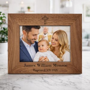 May include: A wooden picture frame with a family photo. The frame has a cross at the top and text at the bottom that reads "James William Monroe Baptized 5/8/2026". The photo shows a baby with parents.