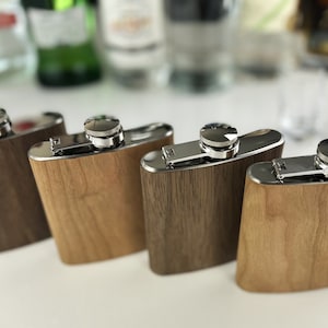 May include: Four stainless steel hip flasks with wood grain patterns. The flasks have a silver colored hinged lid and a silver colored cap.