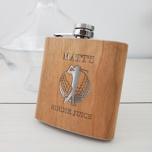 May include: A wooden flask with a silver top and a golfer design. The flask is engraved with the text "Matt's Birdie Juice".