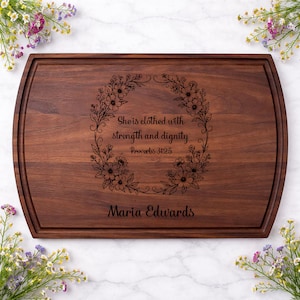 May include: A rectangular wooden cutting board with a dark brown finish. The board features a decorative floral wreath with the text "She is clothed with strength and dignity Proverbs 31:25" and the name "Maria Edwards" engraved in a script font.