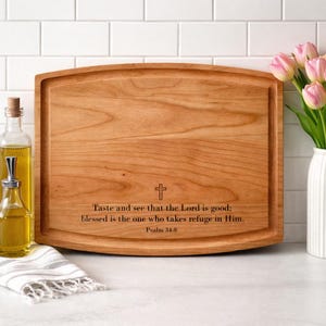 May include: A light brown wooden chopping board with a carved border. The board has the text "Taste and see that the Lord is good: blessed is the one who takes refuge in Him. Psalm 34:8" and a cross symbol.