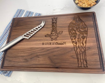 May include: A brown wooden cutting board with a giraffe design engraved on it. The giraffe is shown in two parts, with the head and neck sticking out from the top of the board and the legs and tail extending from the bottom. The text "Did someone say Cheese?!" is engraved below the giraffe's head. A cheese knife is lying on the board.