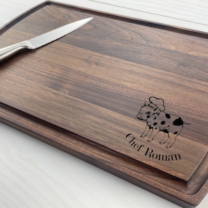 May include: A dark brown wooden chopping board with a raised edge and a pig wearing a chef's hat engraved on it. The text "Chef Roman" is engraved below the pig.