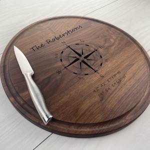 May include: A round wooden chopping board with a compass design and the text "The Robertsons" engraved on it. The board also includes the coordinates "45° 42' 87.99" N and "79° 84' 39.63" W. A silver knife is placed on the board.