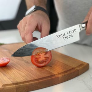 May include: A person is using a silver chef's knife with a wooden handle to cut a red tomato on a wooden chopping board. The knife has the text "Your Logo Here" engraved on the blade.