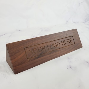 May include: A dark brown wooden desk sign with the engraved text "YOUR LOGO HERE". The sign is rectangular with a triangular base, and the text is centered within a recessed rectangular area.