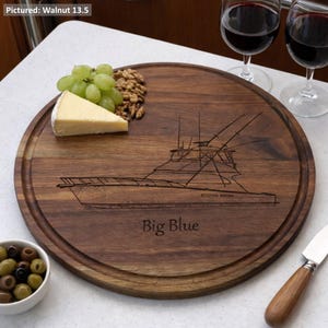 May include: A circular, dark walnut wooden serving board featuring a boat design and the words "Big Blue". The board, with a groove around the edge, holds grapes, cheese, and walnuts. Two glasses of red wine are visible in the background.