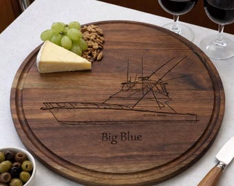 May include: A round, dark walnut wooden serving board with a boat design and the words "Big Blue" engraved on it. The board is surrounded by a groove and is holding grapes, cheese, and walnuts. Two glasses of red wine are in the background.