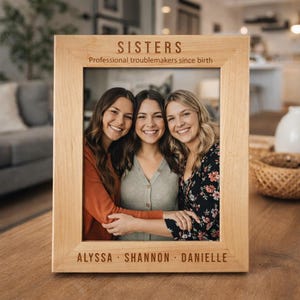 May include: A light-coloured wooden picture frame with the word "SISTERS" at the top and the phrase "Professional troublemakers since birth" below it. The frame holds a photo of three smiling women. The names "ALYSSA", "SHANNON", and "DANIELLE" are at the bottom.