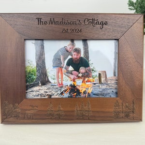 May include: A brown wooden picture frame with a photo of a father and daughter by a campfire. The frame is engraved with the text "The Madison's Cottage Est. 2024" and has a carved design of trees and a moose.