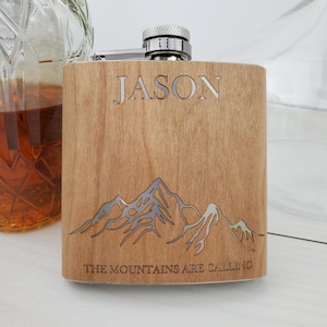 May include: A wooden flask with a stainless steel top. The flask is engraved with the name "JASON" and the text "THE MOUNTAINS ARE CALLING". The flask also features a mountain range design.