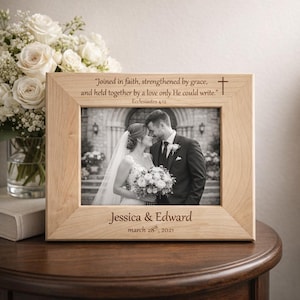 May include: A light-colored wooden picture frame with a black and white wedding photo. The frame has engraved text: "Joined in faith..." and "Jessica & Edward, march 28th, 2021." A bouquet of white flowers is in the background.