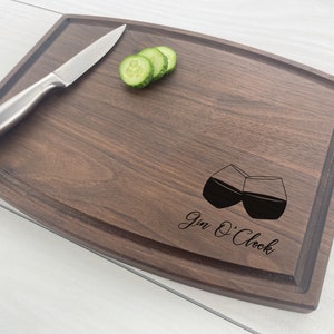 May include: A dark brown wooden cutting board with a rounded edge. The board has a groove around the edge and is engraved with two glasses of gin and the text "Gin O'Clock".