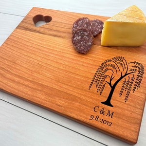 May include: A rectangular wooden cutting board with a heart-shaped cutout and a weeping willow tree design. The board is made of light brown wood and features the initials "C & M" and the date "9.8.2012". Sliced salami and a wedge of cheese are on the board.
