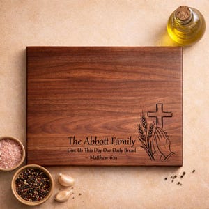 May include: A rectangular wooden chopping board with engraved text and a religious design. The text reads "The Abbott Family Give Us This Day Our Daily Bread - Matthew 6:11." The design includes a cross, wheat, and praying hands. Bowls of spices, garlic, and oil surround the board.