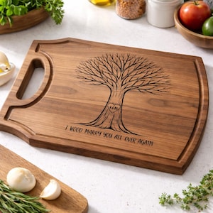 May include: A wooden chopping board with a handle and a tree design. The tree features a heart with initials carved into the trunk. The phrase "I WOOD MARRY YOU ALL OVER AGAIN" is etched below the tree. The board is rectangular.
