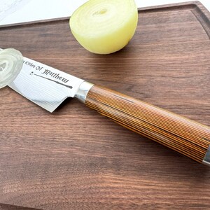Może przedstawiać: Stalowy n&oacute;ż kuchenny z drewnianym uchwytem na drewnianej desce do krojenia. N&oacute;ż ma wz&oacute;r damasceński i jest grawerowany napisem "Kitchen of Matthew".