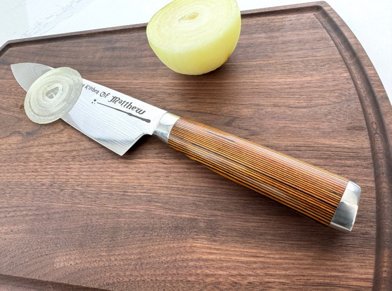 Może przedstawiać: Stalowy n&oacute;ż kuchenny z drewnianym uchwytem na drewnianej desce do krojenia. N&oacute;ż ma wz&oacute;r damasceński i jest grawerowany napisem "Kitchen of Matthew".