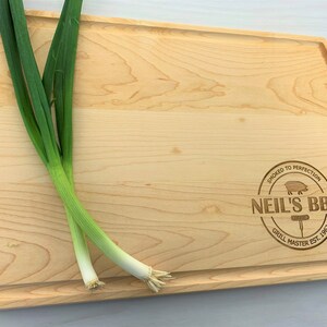 May include: A natural wood cutting board with a rounded edge and a personalized engraved design that reads "Smoked to Perfection Neil's BBQ Grill Master Est. 1962".