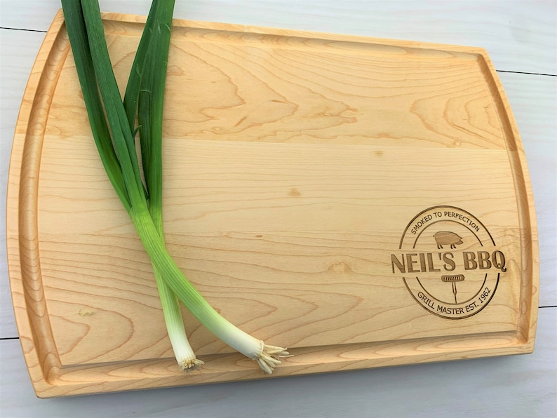 May include: A natural wood cutting board with a rounded edge and a personalized engraved design that reads "Smoked to Perfection Neil's BBQ Grill Master Est. 1962".