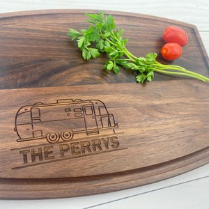 May include: A wooden cutting board with a personalized design featuring a vintage camper and the text "THE PERRYS". The board has a natural wood grain and a curved edge.