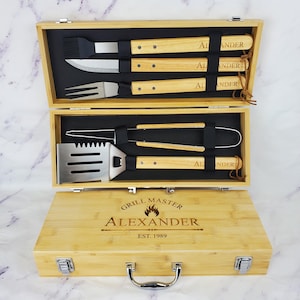May include: A wooden barbecue tool set with stainless steel tools and wooden handles. The set includes a spatula, tongs, knife, fork, and basting brush. The box is engraved with "GRILL MASTER ALEXANDER EST. 1989".