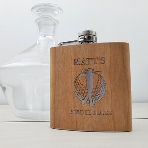 May include: A wooden flask with a silver top and a golfer design. The flask is engraved with the text "Matt's Birdie Juice".