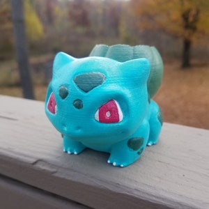 Peut inclure: Un pot de fleur Bulbasaur turquoise avec des yeux rouges et des taches vert foncé. La figurine imprimée en 3D a une ampoule sur le dos et est posée sur une surface en bois. L'arrière-plan est flou, montrant des arbres et du feuillage.