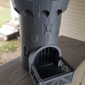Puede incluir: Una torre de dados gris impresa en 3D con forma de torre de castillo. La torre tiene una parte superior almenada, ventanas arqueadas y escudos decorativos. Un pequeño balcón con una puerta enrejada en la base.