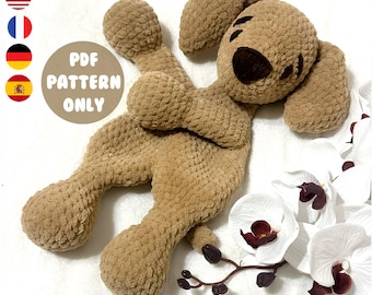 Pode incluir: Um cão de pelúcia de crochê marrom claro, com orelhas caídas e um nariz e olhos marrons escuros. O cão está deitado com os membros estendidos. A imagem inclui o texto "PDF PATTERN ONLY" em um gráfico circular.