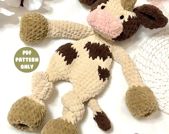 Op de afbeelding: Een handgemaakte gehaakte koeienknuffel in crème, bruin en roze. De koe heeft grote, slappe oren, bruine vlekken en een roze snuit. De tekst "PDF PATTERN ONLY" staat op een groene cirkel. De knuffel ligt op een wit oppervlak.