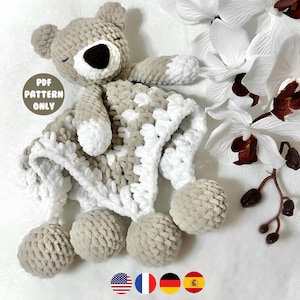 Könnte beinhalten: Eine handgefertigte Häkel-Kuscheldecke in Form eines Bären. Die Decke ist hauptsächlich grau und weiß, mit einem Bärenkopf und vier Pompons. Der Text "PDF PATTERN ONLY" befindet sich in einem Kreis auf der linken Seite des Bildes.