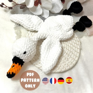Puede incluir: Un cisne blanco de ganchillo con detalles negros y un pico naranja. El cisne está sobre una superficie color crema. La imagen incluye el texto "PDF PATTERN ONLY" en un diseño circular con banderas.