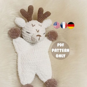 Peut inclure: Jouet en crochet amigurumi en forme de cerf blanc avec des bois et des pompons marrons pour les pieds. Le jouet est couché sur une surface blanche et moelleuse. Le texte "PDF PATTERN ONLY" est visible sur l'image.