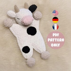 Könnte beinhalten: Ein weißes gehäkeltes Amigurumi-Spielzeug in Form einer Kuh mit schwarzen Flecken. Das Spielzeug hat eine rosa Nase und liegt auf einer beige, flauschigen Oberfläche. Der Text "PDF PATTERN ONLY" ist auf dem Bild zu sehen.