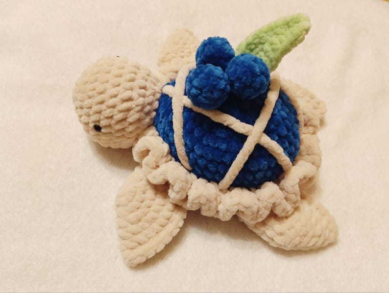 May include: A crocheted plush toy turtle with a blue shell and a green leaf on top. The shell is decorated with blue crocheted blueberries.